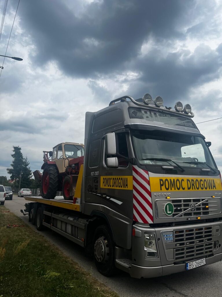 Ciężarówka pomocy drogowej przewozi traktor na lawecie, stojąc na ulicy pod pochmurnym niebem.