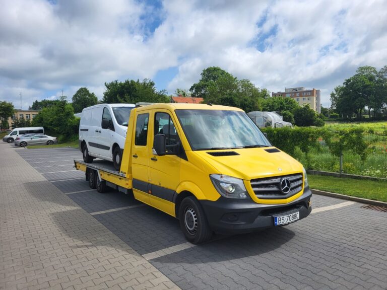 Żółta laweta Mercedes przewożąca biały samochód dostawczy na tle zieleni i budynków w oddali.