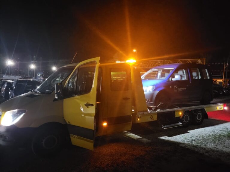 Żółta laweta holująca niebieski samochód nocą z włączonymi światłami ostrzegawczymi.