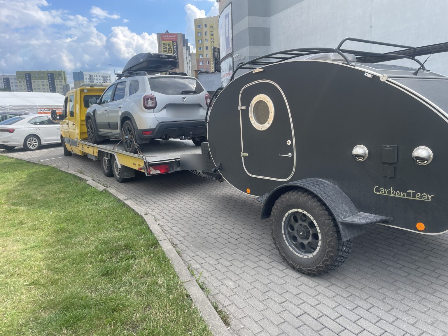 Żółta laweta SKI TRANS wiezie srebrnego SUV-a i ciągnie czarną przyczepę kempingową
